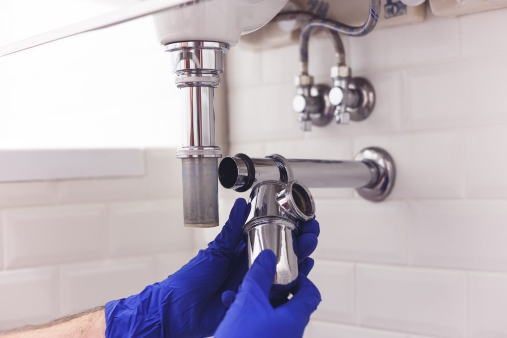 Plumber repairs and maintains chrome siphon under the washbasin. Plumber at work in bathroom, plumbing assemble and install concept "Plumber repairing leaking pipes under kitchen sink in Worcester Park"